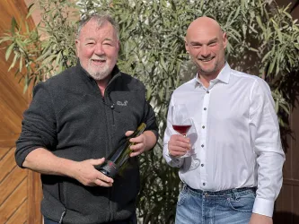 Zwei Männer stehen lächelnd vor einer Holzwand und einer Pflanze. Einer hält eine Flasche Wein, der andere ein Glas mit rotem Getränk.