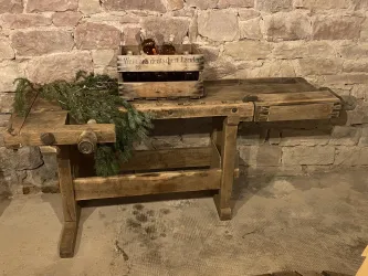 Ein alter Holztisch steht in einem Keller mit einer Steinmauer im Hintergrund. Auf dem Tisch befindet sich eine Kiste mit Flaschen und etwas grünes Zweigwerk.