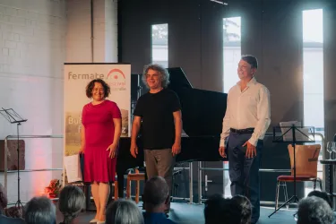 Drei Personen stehen auf einer Bühne und lächeln. Im Hintergrund ist ein Piano und eine entspannte Atmosphäre während einer Veranstaltung zu sehen.