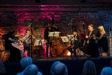 Ein Ensemble mit mehreren Musikern spielt auf einer Bühne. Im Hintergrund ist eine Steinmauer zu sehen, die in dunklen Tönen beleuchtet ist.