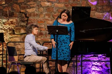 Ein Sänger und ein Pianist treten auf einer Bühne auf. Der Sänger trägt ein blaues Kleid und der Pianist spielt an einem Klavier.