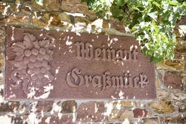 Ein Wirtshausschild mit der Aufschrift „Weingut Grafsmück“. Das Schild ist in den Felsen eingearbeitet und von grünen Pflanzen umgeben.