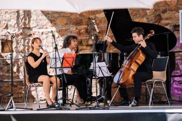 Ein Trio aus Musikern spielt auf einer Bühne. Im Hintergrund ist ein Klavier zu sehen, während die Sängerin und der Cellist auftreten.