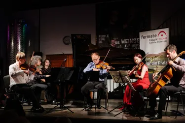 Ein Musikerensemble spielt auf der Bühne. Die Aufführung findet in einem stilvollen Raum mit einem Klavier im Hintergrund statt.