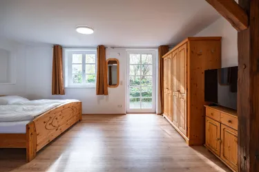 Ein helles Schlafzimmer mit einem Doppelbett aus Holz und einem großen Fenster. Im Raum befinden sich ein Fernseher und ein Schrank aus massivem Holz.