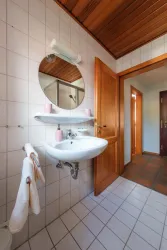 Ein modernes Badezimmer mit einem runden Spiegel und einem Waschbecken. Die Wände sind gefliest und es gibt Holzdetails an der Decke.