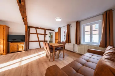 Ein gemütliches Wohnzimmer mit einem braunen Sofa und einem Esstisch. Große Fenster lassen viel Tageslicht herein.