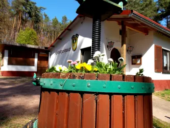 Ein Weinfass mit bunten Blumen steht im Vordergrund. Im Hintergrund sieht man ein gemütliches Gebäude mit Fenstern und einem Wappen.