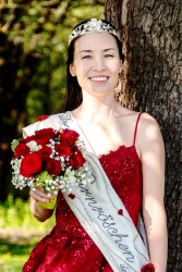 Eine Frau in einem roten Kleid steht neben einem Baum und hält einen Blumenstrauß mit roten Rosen. Sie trägt eine Tiara und ein Schild mit der Aufschrift "Die Schönsten".