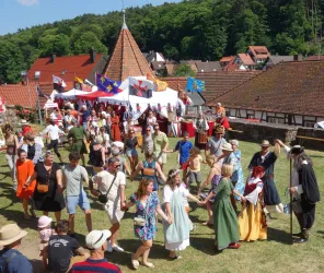 Eine lebhafte Feier im Freien mit Menschen, die tanzen und feiern. Bunte Stände sind im Hintergrund sichtbar, umgeben von einer malerischen Landschaft.