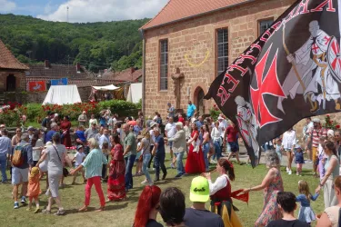 Eine lebhafte Menschenmenge feiert auf einem Fest. Hinter ihnen weht eine große Fahne und historische Gebäude sind im Hintergrund sichtbar.