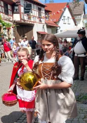 Ein festlicher Straßenumzug mit Kindern in traditionellen Kostümen. Im Vordergrund stehen ein Mädchen in einem roten Umhang und ein weiteres Mädchen, das eine goldene Kugel trägt.