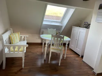 Ein helles Zimmer mit einer schrägen Wand und einer großen Fensterfläche. Es gibt einen kleinen Esstisch, zwei Stühle und eine Bank im skandinavischen Stil.