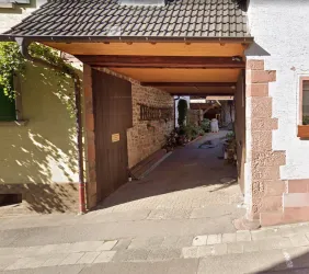 Eine schmale Einfahrt mit einem Holztor und Ziegelwänden. Auf der linken Seite sind Pflanzen und ein gepflasterter Weg sichtbar.
