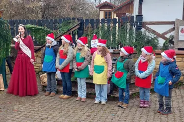 Eine Gruppe von Kindern in Weihnachtsmützen steht in bunten Schürzen vor einer Frau in einem roten Kleid. Die Szene findet draußen statt, umgeben von einer festlichen Atmosphäre.