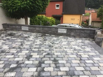 Eine gepflasterte Auffahrt mit grauen Steinen. Im Hintergrund sind Häuser und Sträucher zu sehen.
