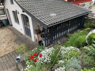 Ein gemütliches Haus mit einem schwarzen Carport und einem blühenden Garten. Der Garten ist bunt mit roten Tulpen und grünen Pflanzen.