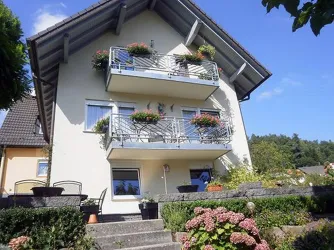 Ein zweistöckiges Haus mit Balkonen, die mit Pflanzen dekoriert sind. Der Himmel ist klar und blau, umgeben von einem grünen Garten.