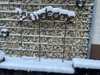 Ein rustikales "Welcome"-Schild aus Metall steht vor einer mit Schnee bedeckten Steinmauer. Einige Herzen aus Metall schmücken die Wand im Hintergrund.