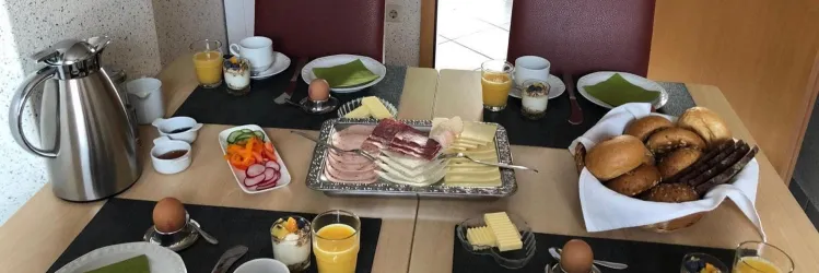 Ein reichhaltiges Frühstück mit frischen Brötchen, Wurst, Käse und Obst. Dazu gibt es Eier, Joghurt und verschiedene Getränke.