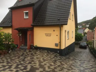 Ein modernes, zweifarbige Haus mit einer gepflasterten Einfahrt. Die Umgebung ist grün und zeigt einige angrenzende Gebäude.