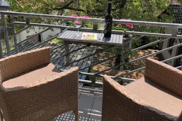 Ein gemütlicher Balkon mit zwei Stühlen und einem kleinen Tisch. Auf dem Tisch steht eine Flasche Wein und einige Blumen sind dekoriert.