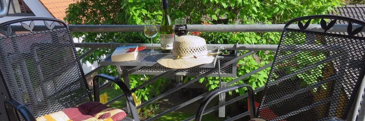 Ein Balkon mit zwei schwarzen Stühlen und einem kleinen Tisch. Auf dem Tisch liegen ein Hut, ein Buch und ein Glas Wein, umgeben von grünem Pflanzen.