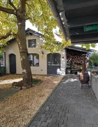 Ein charmantes Haus mit einem großen Baum und einem angelegten Weg. Im Hintergrund sind Holzstapel und eine Feuerstelle sichtbar.