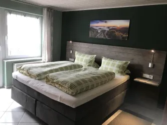 Ein gemütliches Schlafzimmer mit einem großen Doppelbett und zwei Kissen. An der Wand hängt ein Bild einer Landschaft und es gibt ein Fenster mit viel Tageslicht.