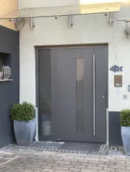 Eine moderne Haustür in Grau mit einem großen Fenster. Vor der Tür stehen zwei große Pflanzkübel mit grünen Pflanzen.