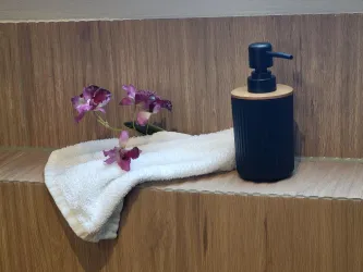 Eine schwarze Seifendose steht auf einer Ablage mit Fliesen in Holzoptik neben einem weißen Handtuch. Daneben sind lila Blumen in einer kleinen Vase platziert.