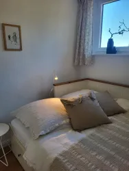 Ein gemütliches Schlafzimmer mit einem Doppelbett und Kissen. Neben dem Bett steht ein kleiner Tisch und ein Fenster mit Blick nach draußen.