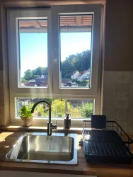 Eine helle Küche mit einem Edelstahlspülbecken und einem Ausblick auf grüne Hügel. Draußen sind Häuser sichtbar und die Sonne scheint durch das Fenster.