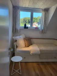 Ein gemütliches Schlafzimmer mit einem Tagesbett und einem kleinen Tisch. Durch das Fenster fällt helles Licht auf die hellen Wände und den Holzboden.