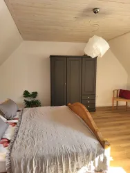 Ein helles Schlafzimmer mit einem großen Bett und einem stilvollen Schrank. Es gibt einen gemütlichen Stuhl und eine Pflanze im Raum.