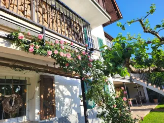 Ein charmantes Haus mit einem Balkon, der mit rosa Rosen geschmückt ist. Im Vordergrund sind grüne Pflanzen und ein klarer blauer Himmel zu sehen.