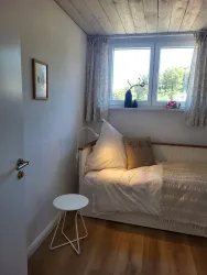 Ein helles Schlafzimmer mit einem Bett und einem kleinen Tisch. Die Fenster bieten Blick auf das grüne Außenareal.