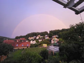 Ein Regenbogen spannt sich über ein malerisches Dorf mit bunten Häusern. Die Landschaft ist grün und idyllisch, umgeben von sanften Hügeln.