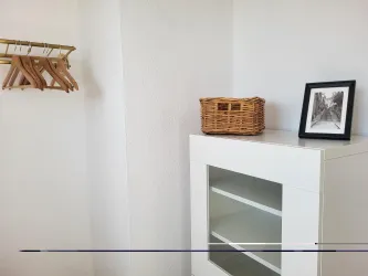 Ein heller Raum mit einem weißen Möbelstück und einer Korb. An der Wand hängen Holzkleiderbügel und ein gerahmtes Bild steht auf dem Regal.