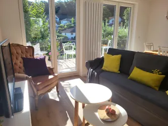 Ein helles Wohnzimmer mit einem grauen Sofa und einem beigen Sessel. Der Blick auf einen Balkon mit Pflanzen und Sonnenschein sorgt für eine einladende Atmosphäre.