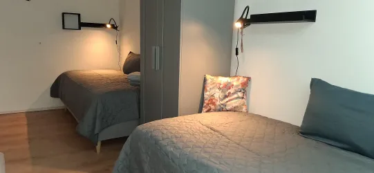 Ein einfaches Zimmer mit zwei Einzelbetten und einer modernen Lampe. Die Wände sind hell und die Einrichtung ist minimalistisch.