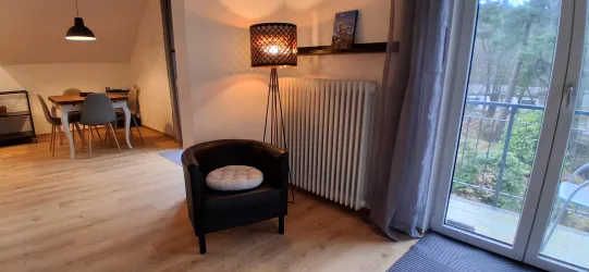 Ein gemütlicher Wohnraum mit einem schwarzen Sessel und einer Stehlampe. Die große Fenstertür bietet einen Blick ins Freie, während der Boden aus Holz eine warme Atmosphäre schafft.