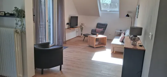 Ein gemütliches Wohnzimmer mit Holzfußboden und viel Licht. Es gibt einen Fernseher, einen Sessel und eine kleine Sitzecke mit einem Tisch.