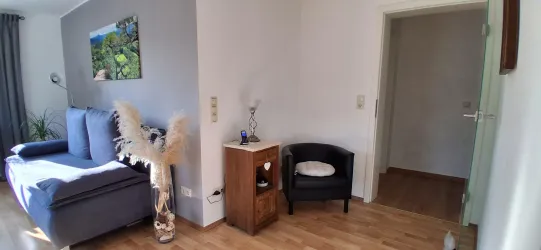 Ein gemütliches Wohnzimmer mit einem grauen Sofa und Pflanzen. An der Wand hängt ein Bild der Natur, und es gibt einen kleinen Tisch mit einem Stuhl.