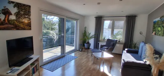 Ein modernes Wohnzimmer mit einem gemütlichen Sofa und einem Fernseher. Große Fenster lassen viel Licht herein und bieten einen Blick auf Pflanzen im Freien.