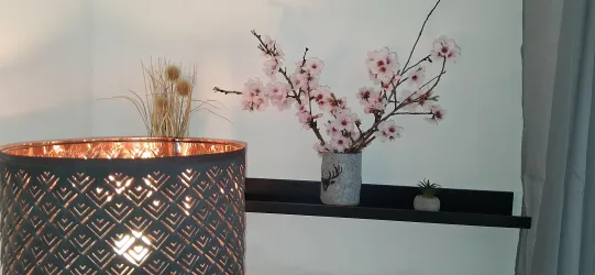 Eine stilvolle Lampe mit einem dekorativen Schirm steht neben einer Wand. Im Hintergrund sind Blütenzweige in einer Vase zu sehen, die dem Raum eine gemütliche Atmosphäre verleihen.