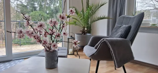 Ein gemütlicher Raum mit einem modernen Sessel und einer Blumenvase auf dem Tisch. Im Hintergrund sind Pflanzen und große Fenster, die Tageslicht hereinlassen.