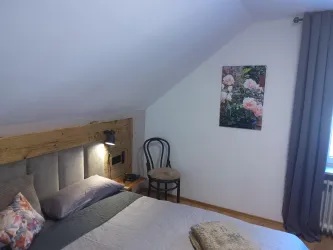 Ein gemütliches Schlafzimmer mit einem schlichten Bett und einem Stuhl. An der Wand befindet sich ein Blumenbild, was eine beruhigende Atmosphäre schafft.