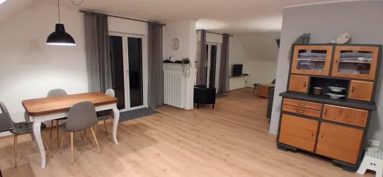 Ein modernes Wohnzimmer mit einem Esstisch und Stühlen. Die Räume sind hell und gemütlich eingerichtet, mit Holzfußboden und großen Fenstern.