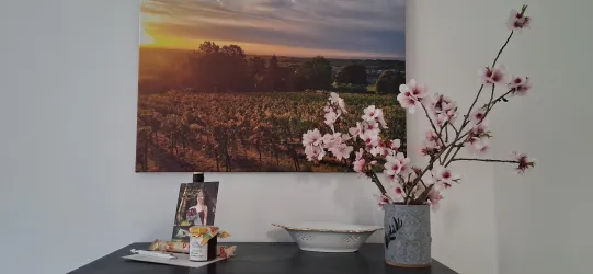 Ein gemütlicher Tisch mit einer Vase voller blühender Zweige und einem schönen Foto. Im Hintergrund hängt ein Bild von einer Landschaft bei Sonnenuntergang.